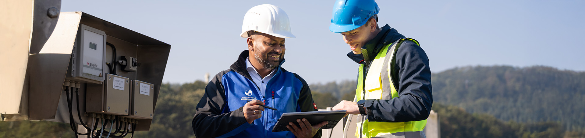 2 Personen mit Weste und Helm an stehen vor einer technischen Anlage, schauen auf ein Tablet und interagieren miteinander.