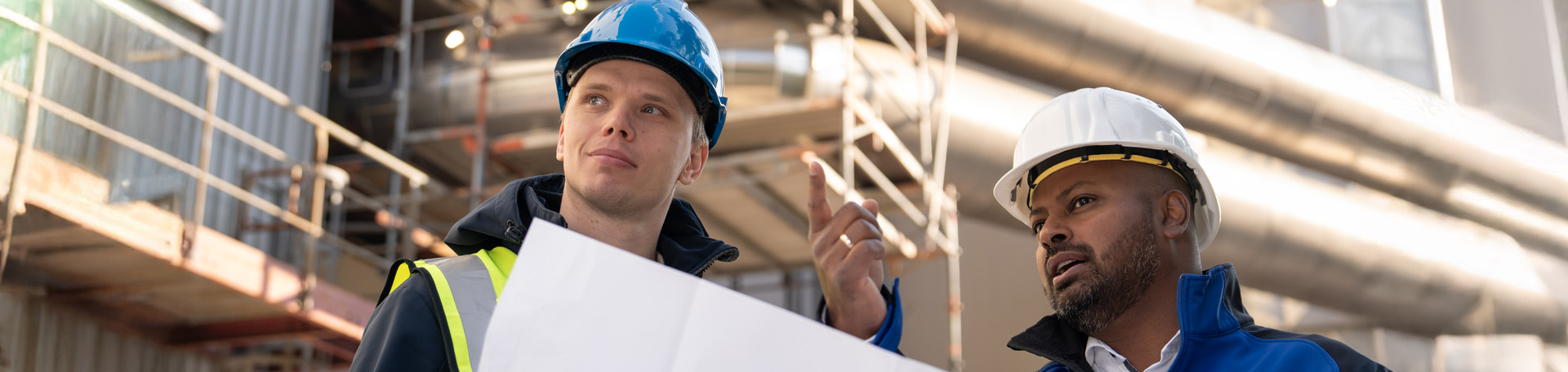 2 Ingenieure mit Helm und Weste an stehen vor einer großen Anlage, halten einen Plan in der Hand, schauen in die Ferne und sprechen miteinander.