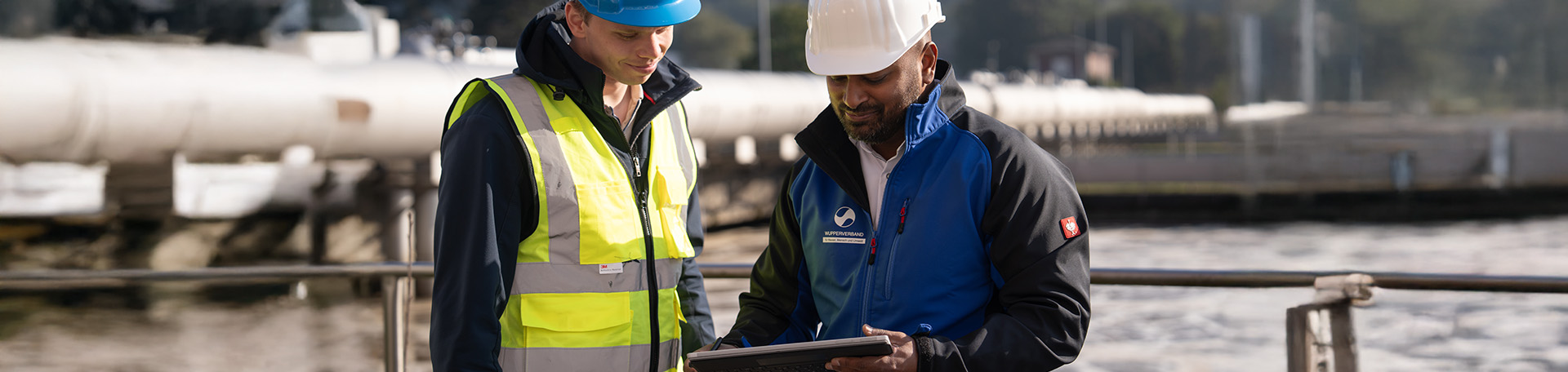 2 Personen mit gelber Weste und Helm an stehen am Rande eines Belebungsbeckens einer Kläranlage und schauen auf ein Tablet. Im Hintergrund sind dicke Rohrleitungen zu sehen.