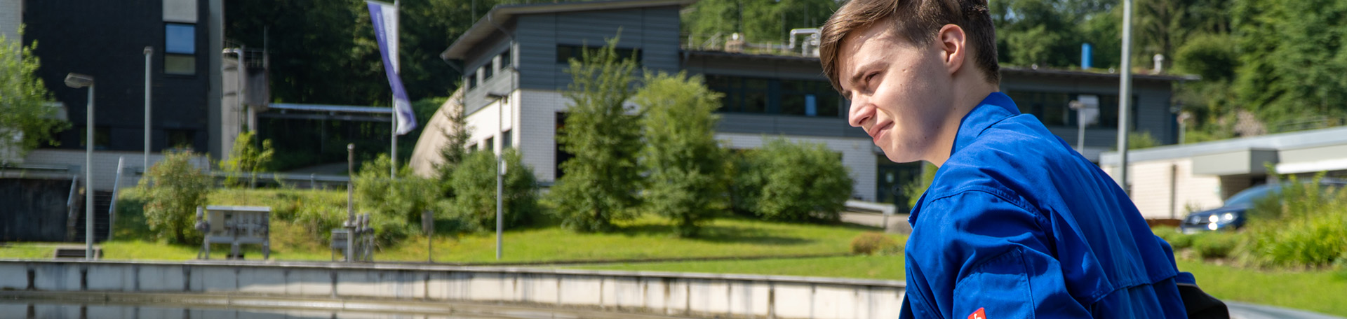 Ein junger Mann steht bei strahlendem Sonnenschein in blauer Arbeitskleidung am Rande eines runden Klärbeckens. Im Hintergrund ist eine grüne Wiese, Bäume und einige Gebäude zu sehen.
