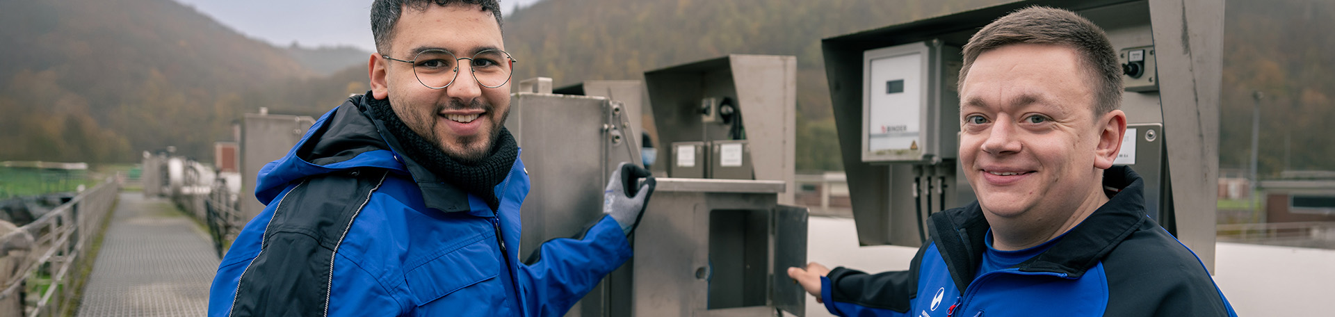 2 Personen in Arbeitskleidung stehen auf dem Steg eines Klärbeckens an einer technischen Anlage und schauen lächelnd in die Kamera.